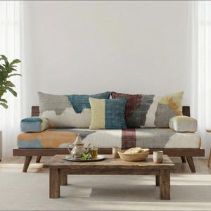 May include: A living room scene with a wooden sofa and patterned cushions, a coffee table, and a potted plant. A side table with books is also present. Sheer white curtains frame the windows.
