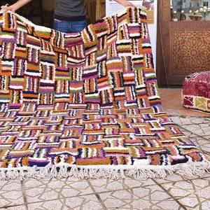 May include: A colorful Moroccan rug with a geometric pattern in shades of orange, purple, white, and black. The rug has a fringe border and is being held up by a person.