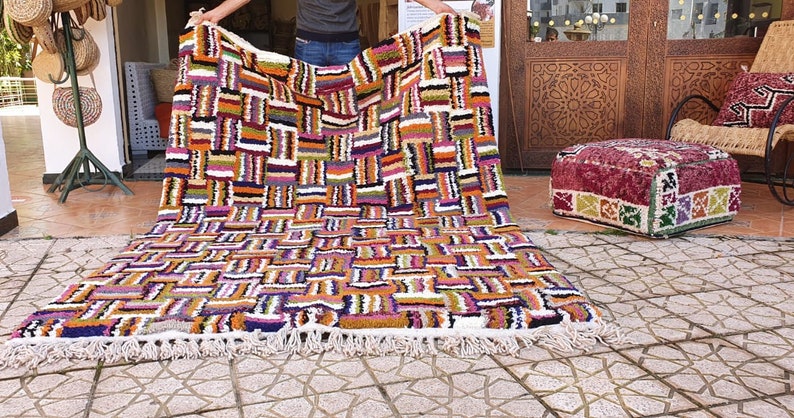 May include: A colorful Moroccan rug with a geometric pattern in shades of orange, purple, white, and black. The rug has a fringe border and is being held up by a person.