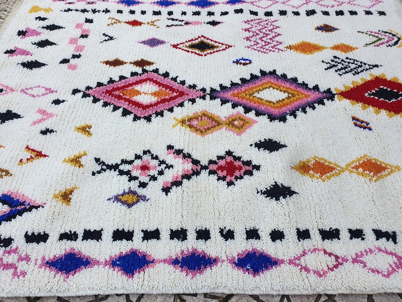 May include: A white rug with a colorful geometric pattern. The pattern features diamonds, triangles, and other shapes in shades of pink, red, blue, yellow, and black.