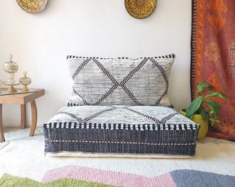 Pode incluir: Um almofada de chão com padrão preto e branco com uma almofada combinando. O almofada é feito de tecido tecido e tem um design acolchoado. O almofada está colocado sobre um tapete branco com um design rosa e verde.