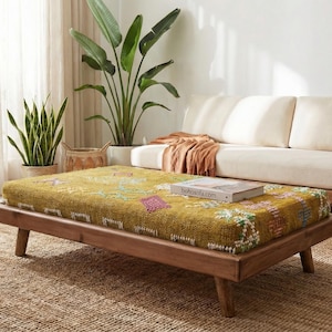 May include: A living room scene featuring a low, wooden-framed ottoman with a patterned yellow cushion. A cream-coloured sofa and various potted plants are in the background. A woven rug covers the floor.
