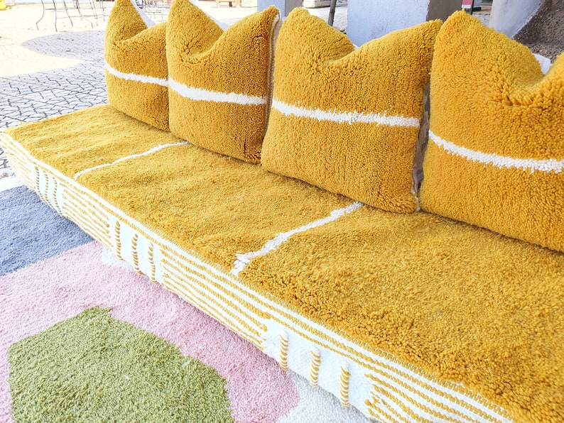 May include: A yellow tufted sofa with white stripes along the edges and four yellow tufted pillows with white stripes. The sofa is on a multi-colored rug.