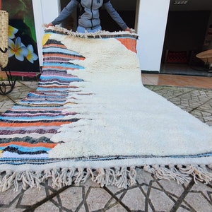 Op de afbeelding: Een wit en kleurrijk Marokkaans tapijt met franjes. Het tapijt heeft een witte achtergrond met een kleurrijk geometrisch patroon aan de ene kant. Het tapijt wordt door een persoon omhoog gehouden.