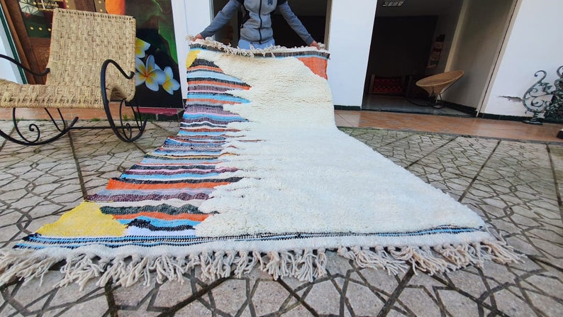 Op de afbeelding: Een wit en kleurrijk Marokkaans tapijt met franjes. Het tapijt heeft een witte achtergrond met een kleurrijk geometrisch patroon aan de ene kant. Het tapijt wordt door een persoon omhoog gehouden.