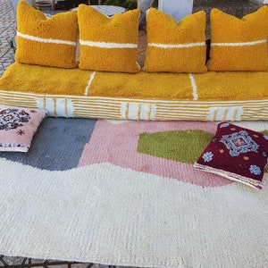 May include: A yellow and white striped pouf with four yellow throw pillows with white stripes. The pouf is sitting on a white rug with pink, green, and gray geometric shapes. Two smaller pillows with embroidered designs are on the rug.