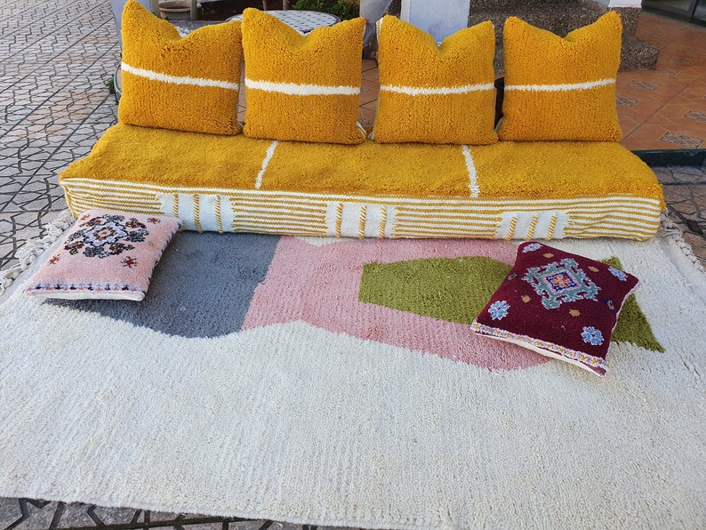 May include: A yellow and white striped pouf with four yellow throw pillows with white stripes. The pouf is sitting on a white rug with pink, green, and gray geometric shapes. Two smaller pillows with embroidered designs are on the rug.