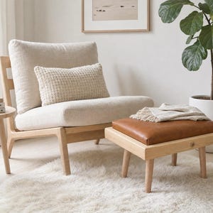 May include: A light-colored wooden lounge chair with a cream-colored upholstered seat and back cushion, accompanied by a matching rectangular ottoman featuring a brown leather-like top cushion and a small cream-colored throw blanket on top.