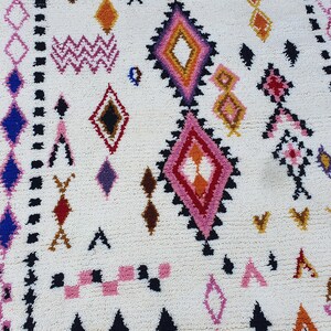 May include: A white rug with a geometric pattern in shades of pink, blue, red, orange, and brown. The rug has a diamond pattern with a border of black and white.