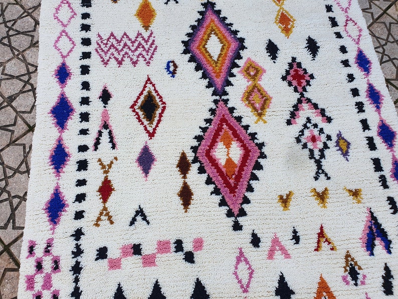 May include: A white rug with a geometric pattern in shades of pink, blue, red, orange, and brown. The rug has a diamond pattern with a border of black and white.
