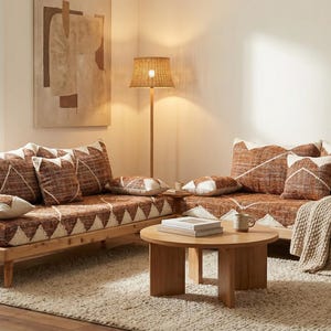 Puede incluir: Un salón acogedor con dos sofás de madera adornados con cojines estampados y una mesa de centro a juego. Una lámpara tejida y una gran planta contribuyen al ambiente cálido. Los sofás presentan un patrón geométrico marrón y blanco.