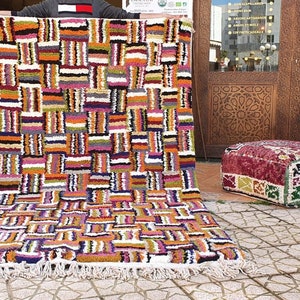 May include: A colorful Moroccan rug with a geometric pattern in shades of orange, pink, white, and black. The rug has a fringe border and is laid out on a tiled floor.