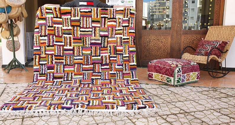 May include: A colorful Moroccan rug with a geometric pattern in shades of orange, pink, white, and black. The rug has a fringe border and is laid out on a tiled floor.