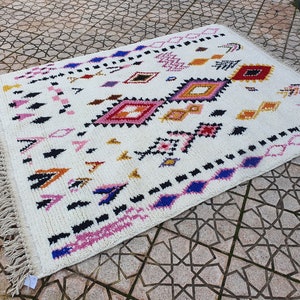 May include: A white rug with a geometric pattern in shades of pink, blue, yellow, and black. The rug has a fringe border.