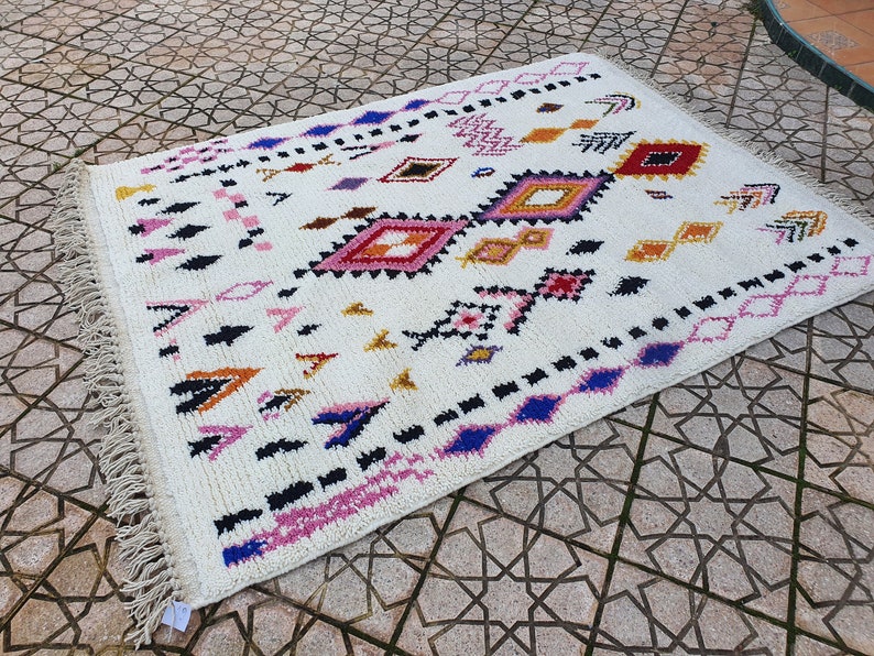May include: A white rug with a geometric pattern in shades of pink, blue, yellow, and black. The rug has a fringe border.