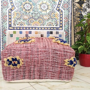 May include: A red and white woven pouf with blue and yellow accents. The pouf is sitting in front of a tiled wall with a Moroccan design.