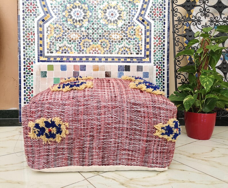 May include: A red and white woven pouf with blue and yellow accents. The pouf is sitting in front of a tiled wall with a Moroccan design.