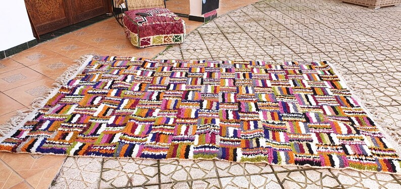 May include: A colorful Moroccan rug with a geometric pattern in shades of orange, pink, purple, green, and white. The rug has a white background and is made of wool.
