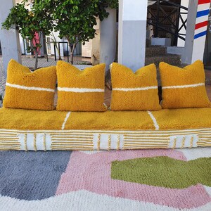 May include: A yellow tufted sofa with white stripes and four matching pillows. The sofa is sitting on a white rug with pink, gray, and green geometric shapes.