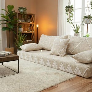 May include: A living room with a low beige sofa with geometric patterns and matching cushions. A dark wood coffee table sits on a cream rug, surrounded by potted plants and hanging greenery. The room has a warm, inviting atmosphere, with a wooden floor.