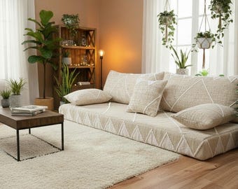 May include: A living room with a low beige sofa with geometric patterns and matching cushions. A dark wood coffee table sits on a cream rug, surrounded by potted plants and hanging greenery. The room has a warm, inviting atmosphere, with a wooden floor.