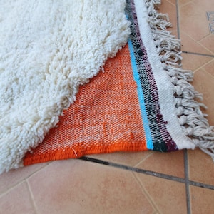 Op de afbeelding: Een wit, pluche tapijt met een franje rand is gedeeltelijk gevouwen over een kleurrijk geweven tapijt met oranje, blauwe, groene en witte strepen. Het geweven tapijt heeft een franje rand.