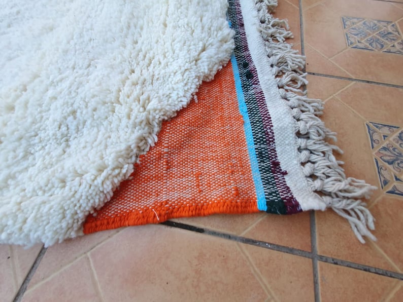 Op de afbeelding: Een wit, pluche tapijt met een franje rand is gedeeltelijk gevouwen over een kleurrijk geweven tapijt met oranje, blauwe, groene en witte strepen. Het geweven tapijt heeft een franje rand.