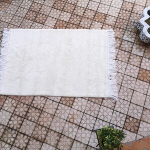 K&ouml;nnte beinhalten: Ein wei&szlig;er, flauschiger Teppich mit Fransen an allen Seiten. Der Teppich liegt auf einem gefliesten Boden mit einem geometrischen Muster.