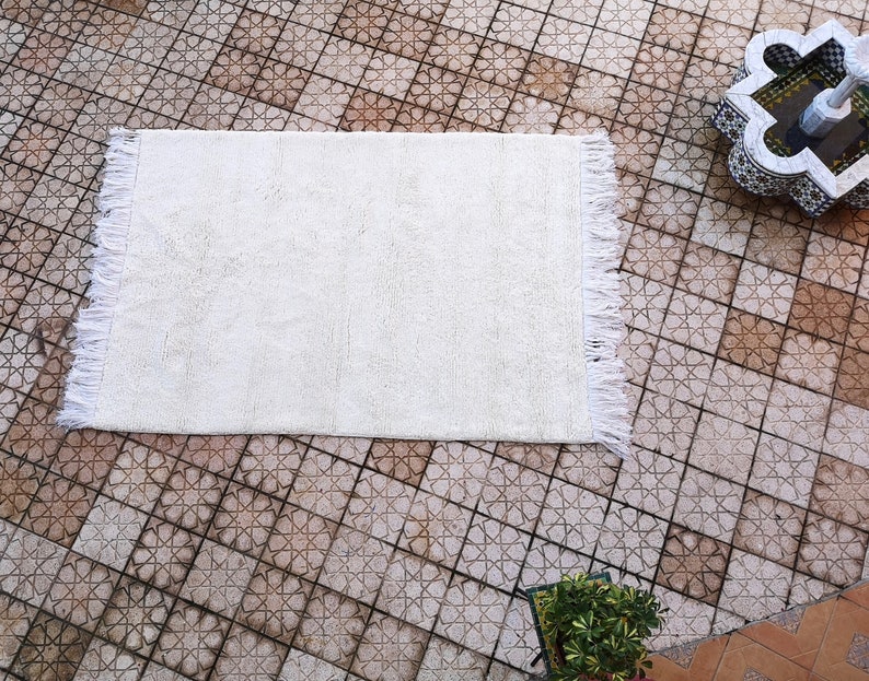 K&ouml;nnte beinhalten: Ein wei&szlig;er, flauschiger Teppich mit Fransen an allen Seiten. Der Teppich liegt auf einem gefliesten Boden mit einem geometrischen Muster.