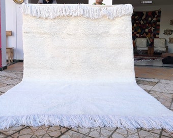 May include: A white, handwoven rug with a fringe border. The rug is folded in half and laid out on a tiled floor.
