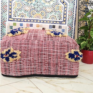 May include: A red and white woven pouf with blue and yellow floral accents. The pouf is sitting on a tiled floor.
