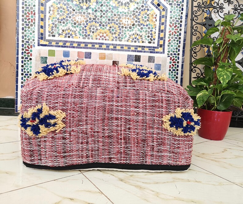 May include: A red and white woven pouf with blue and yellow floral accents. The pouf is sitting on a tiled floor.