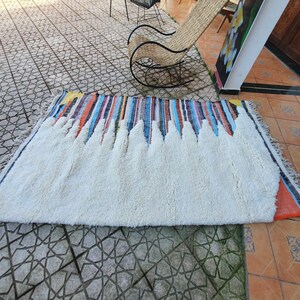 Op de afbeelding: Een wit, pluche vloerkleed met een kleurrijke gestreepte rand. Het vloerkleed ligt op een betegelde vloer.