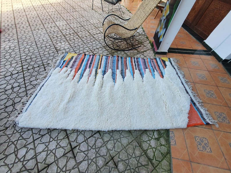 Op de afbeelding: Een wit, pluche vloerkleed met een kleurrijke gestreepte rand. Het vloerkleed ligt op een betegelde vloer.