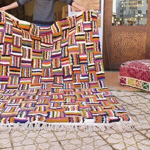 May include: A colorful Moroccan rug with a geometric pattern in shades of orange, purple, pink, and black. The rug has a white fringe border and is laid out on a tiled floor.