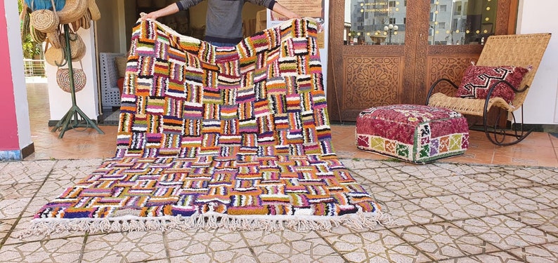 May include: A colorful Moroccan rug with a geometric pattern in shades of orange, purple, pink, and black. The rug has a white fringe border and is laid out on a tiled floor.