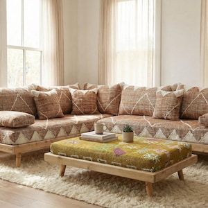 Könnte beinhalten: Ein Ecksofa mit hellem Holzrahmen und gemusterten Kissen in Braun- und Cremetönen. Ein Hocker mit floralem Design steht vor dem Sofa. Eine hohe gewebte Lampe und eine Topfpflanze vervollständigen das Wohnzimmer.