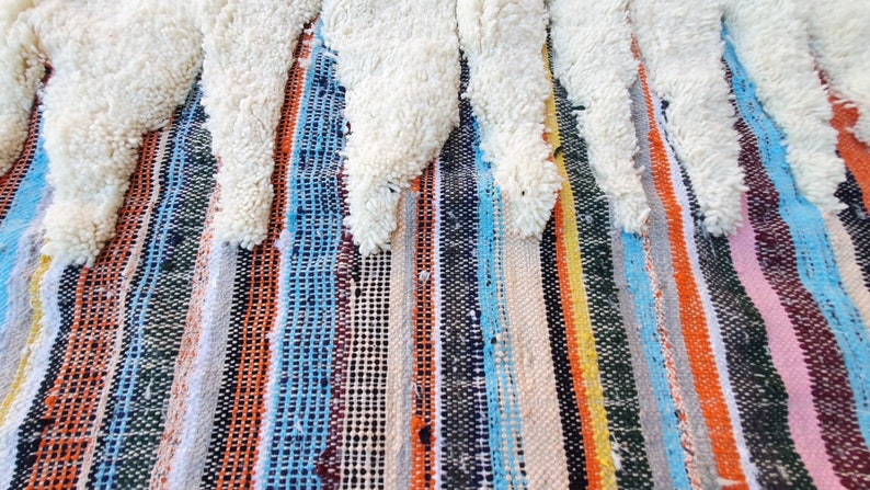 Op de afbeelding: Een kleurrijk gestreept tapijt met een witte, pluche franje. Het tapijt heeft een verscheidenheid aan kleuren, waaronder blauw, oranje, geel en roze.