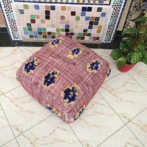 May include: A square, burgundy-colored pouf with a woven texture and a fringe border. The pouf has a floral pattern with blue and yellow accents.
