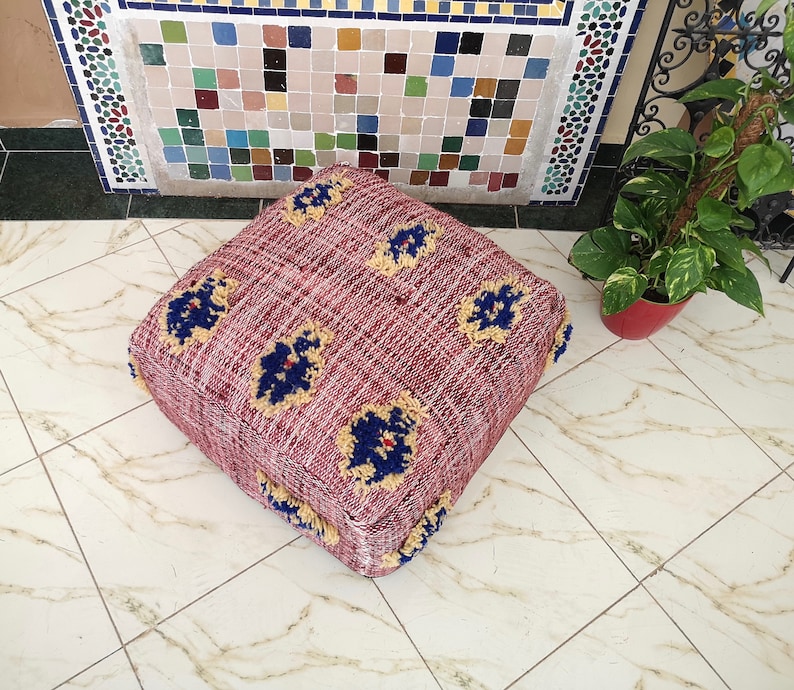 May include: A square, burgundy-colored pouf with a woven texture and a fringe border. The pouf has a floral pattern with blue and yellow accents.