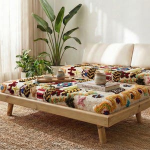 May include: A light wood frame sofa and coffee table set with colorful patterned cushions. The cushions feature a mix of red, blue, yellow, and green designs. The set is placed on a woven jute rug, with plants and sheer curtains in the background.