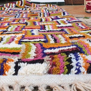 May include: A colorful Moroccan rug with a geometric pattern in shades of orange, pink, green, blue, and white. The rug has a fringe border and is made of wool.