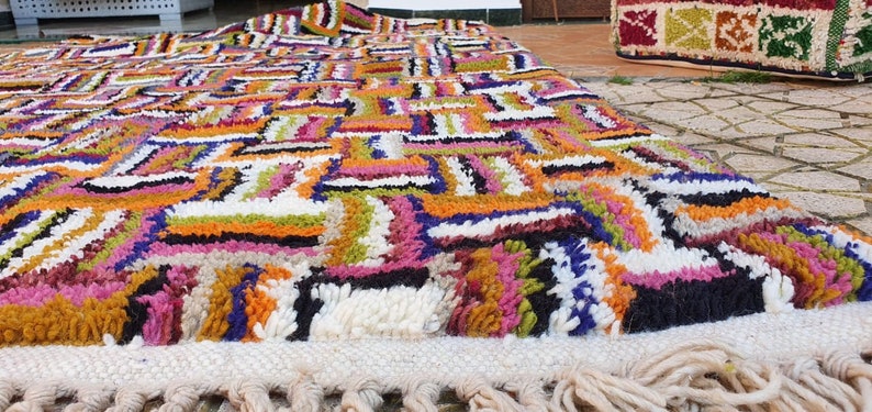 May include: A colorful Moroccan rug with a geometric pattern in shades of orange, pink, green, blue, and white. The rug has a fringe border and is made of wool.