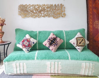 May include: A green, plush, floor cushion with a white trim and three embroidered pillows. The pillows have a pink, white, and brown geometric pattern. The cushion is in front of a white wall with a gold metal wall hanging.