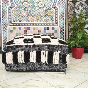May include: Black and white woven pouf with a checkered pattern and fringe detail. The pouf is sitting on a tiled floor.