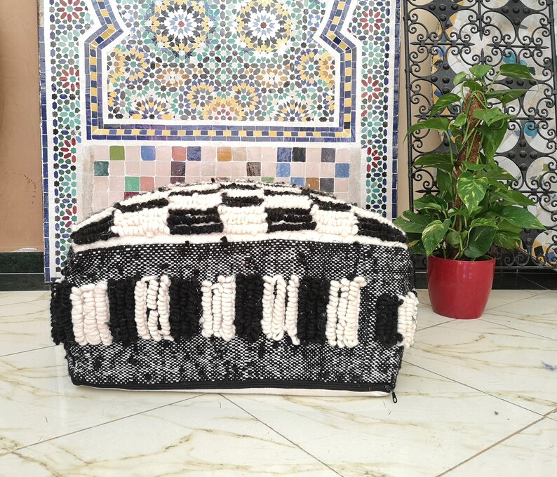 May include: Black and white woven pouf with a checkered pattern and fringe detail. The pouf is sitting on a tiled floor.