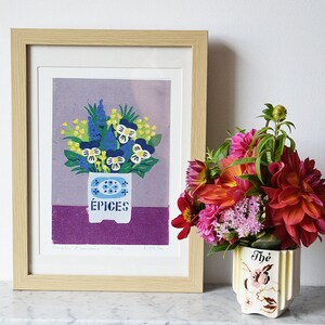 May include: A framed print of a floral arrangement in a white spice container with the word "&Eacute;PICES". The print is in a light wood frame. To the right is a vase with a bouquet of colorful flowers.