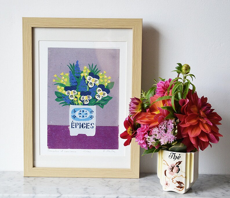 May include: A framed print of a floral arrangement in a white spice container with the word "&Eacute;PICES". The print is in a light wood frame. To the right is a vase with a bouquet of colorful flowers.