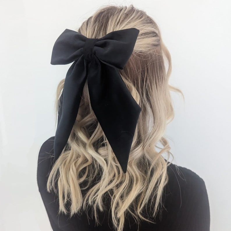 Puede incluir: Un lazo para el pelo grande y de color negro s&oacute;lido con largas colas terminadas en punta. El lazo est&aacute; confeccionado con una tela lisa y mate, y se encuentra sujeto sobre una melena rubia y ondulada recogida parcialmente hacia atr&aacute;s.