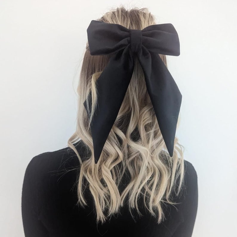 Puede incluir: Un lazo para el pelo grande de color negro s&oacute;lido con extremos largos y puntiagudos. El lazo est&aacute; confeccionado con una tela lisa y mate y se asegura en el centro con un nudo de tela a juego. El dise&ntilde;o presenta una forma estructurada.